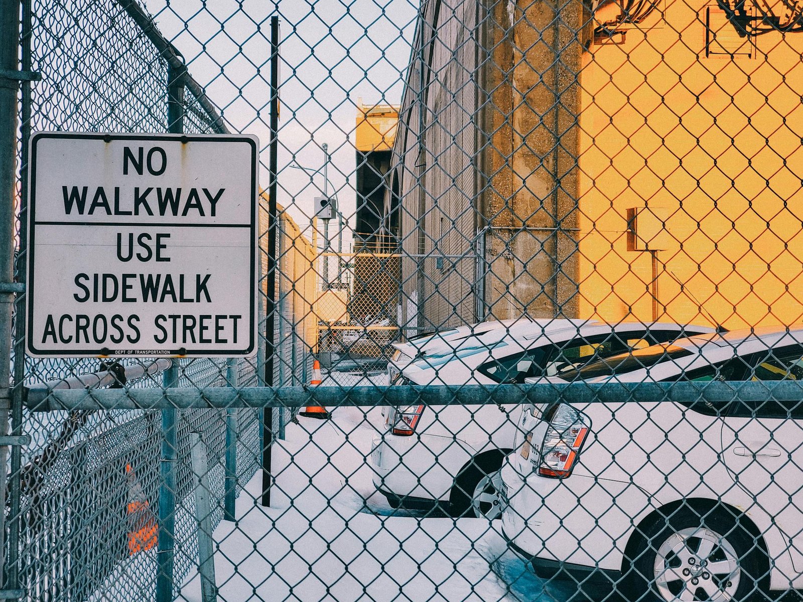 Chain-link fence with warning sign and parked cars in snowy urban setting.